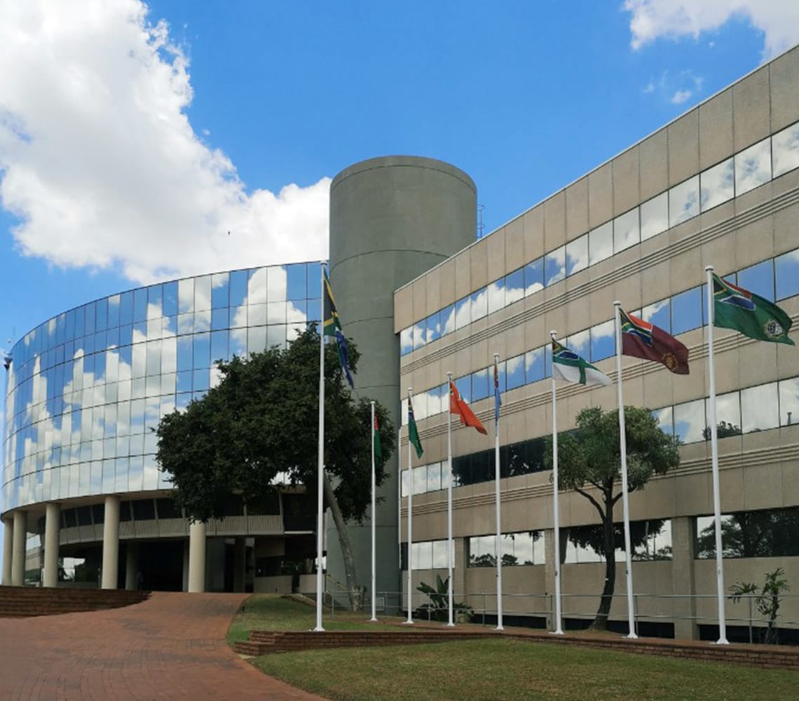 A modern glass and concrete building with a cylindrical section, surrounded by flagpoles displaying various national flags, under a partly cloudy sky.